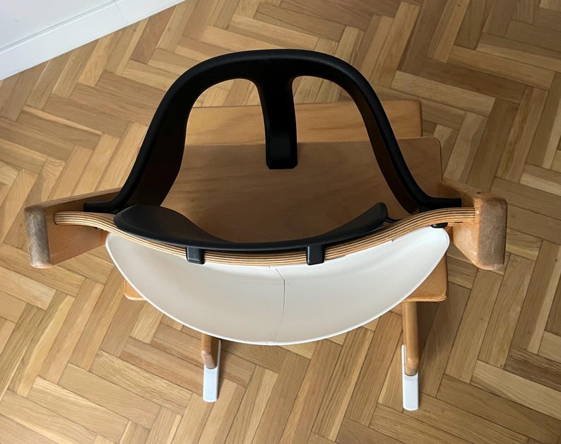 May include: A modern, minimalist rocking chair with a black backrest and seat, and a white, curved base. The chair has wooden side supports and is designed for comfort and style. The chair is set on a wooden surface.