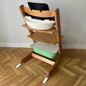 May include: A wooden high chair with a black backrest, a white tray, and a green container. The chair has a natural wood finish and a sturdy design, suitable for a child. The chair is designed for safety and comfort.