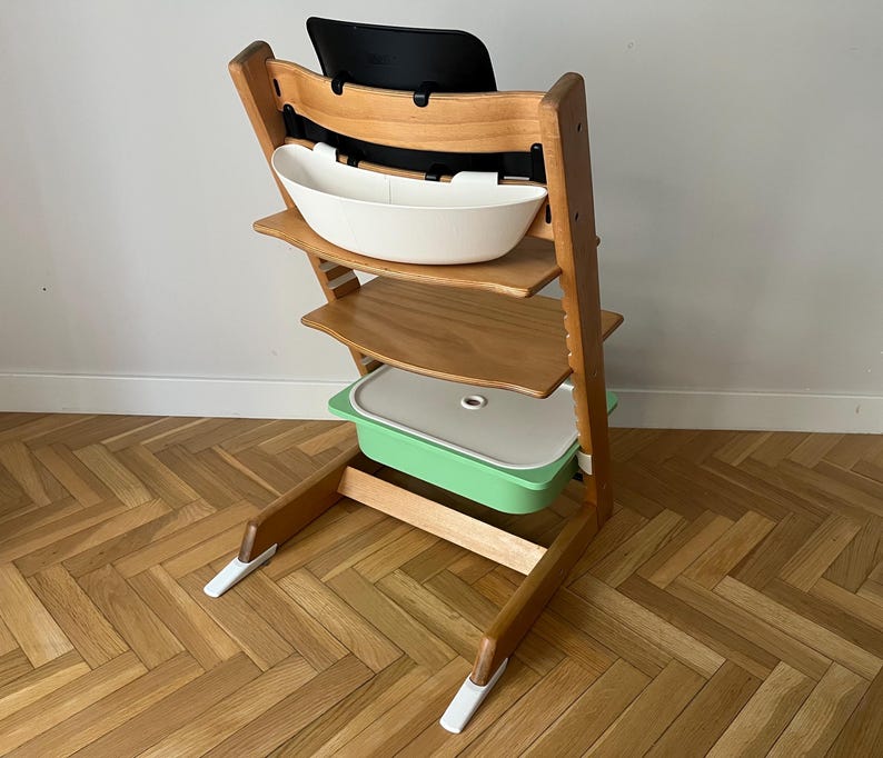 May include: A wooden high chair with a black backrest, a white tray, and a green container. The chair has a natural wood finish and a sturdy design, suitable for a child. The chair is designed for safety and comfort.