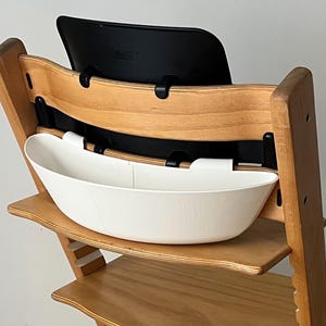 May include: A wooden high chair with a black backrest and a white, oval-shaped tray. The chair has a natural wood finish and is designed for a child. The tray is attached to the front of the chair.