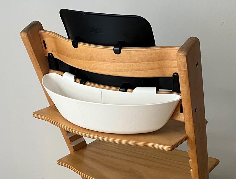May include: A wooden high chair with a black backrest and a white, oval-shaped tray. The chair has a natural wood finish and is designed for a child. The tray is attached to the front of the chair.