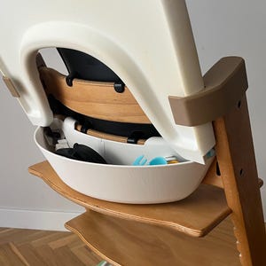 May include: A wooden high chair with a white seat and a white storage bowl. The chair has a brown frame and a beige backrest. The bowl contains blue and black items. The chair is designed for a baby or toddler.