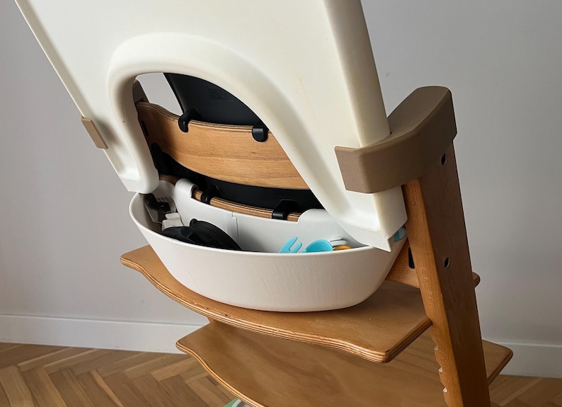 May include: A wooden high chair with a white seat and a white storage bowl. The chair has a brown frame and a beige backrest. The bowl contains blue and black items. The chair is designed for a baby or toddler.