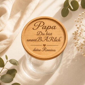 May include: A clear glass jar with a round wooden lid featuring the engraved German text Papa Du bist unentB&Auml;Rlich with a small heart symbol and the name deine Romina. The lid has a natural light brown wood grain finish and circular texture.