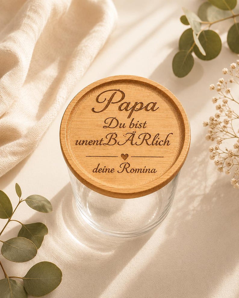 May include: A clear glass jar with a round wooden lid featuring the engraved German text Papa Du bist unentB&Auml;Rlich with a small heart symbol and the name deine Romina. The lid has a natural light brown wood grain finish and circular texture.