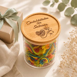 May include: A clear glass jar filled with colorful, multi-colored gummy candy strings. The jar features a round, light-colored wooden lid engraved with the text "Geduldsf&auml;den f&uuml;r Sabrina" and a graphic illustration of a heart-shaped yarn ball.