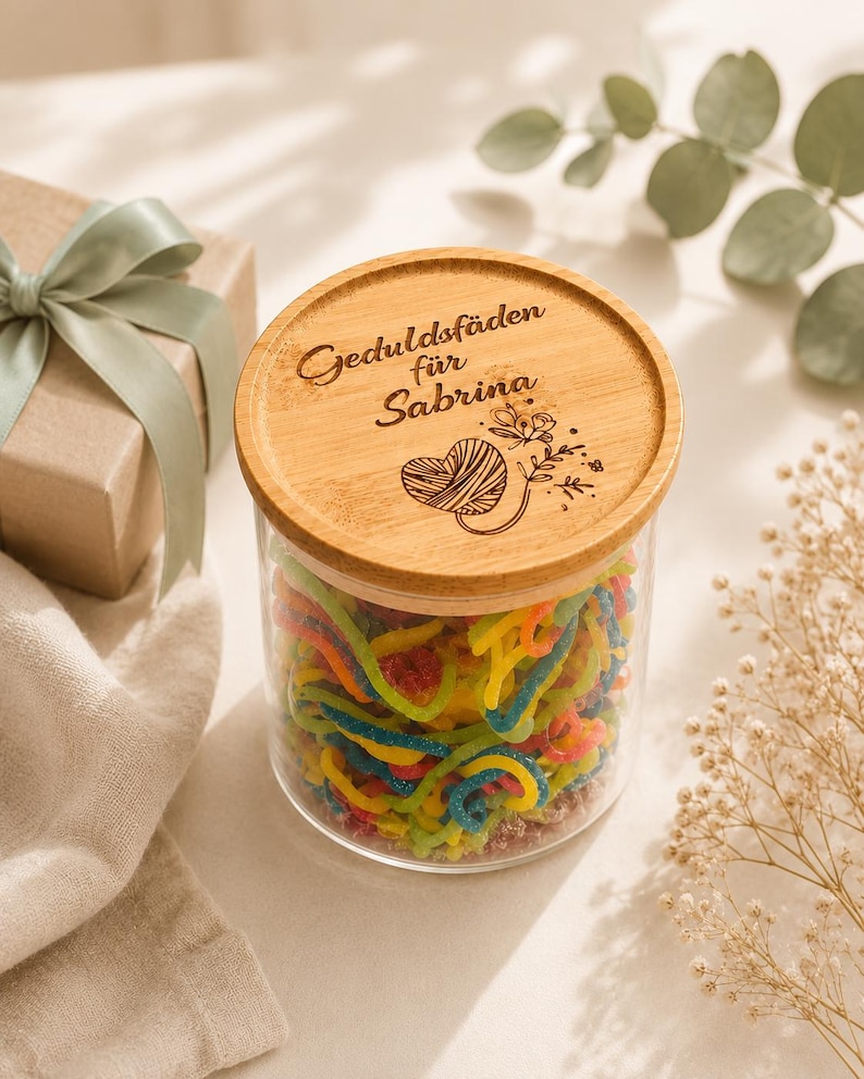 May include: A clear glass jar filled with colorful, multi-colored gummy candy strings. The jar features a round, light-colored wooden lid engraved with the text "Geduldsf&auml;den f&uuml;r Sabrina" and a graphic illustration of a heart-shaped yarn ball.