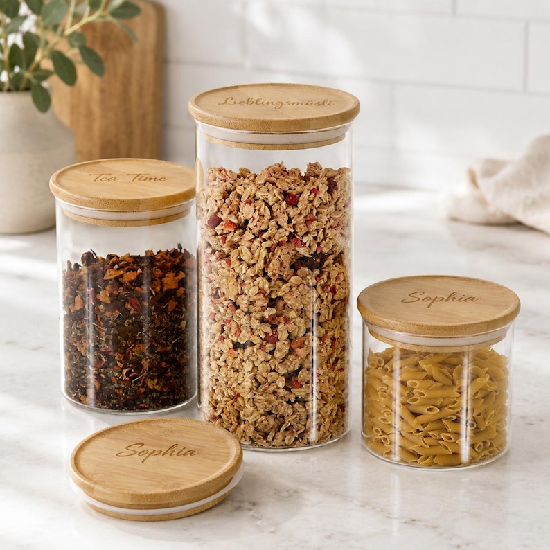 May include: Three clear glass storage canisters with round bamboo lids. The lids are engraved with the text Tea Time, Lieblingsm&uuml;sli, and Sophia. One lid is placed flat on the surface. The canisters contain dried tea, granola, and pasta.