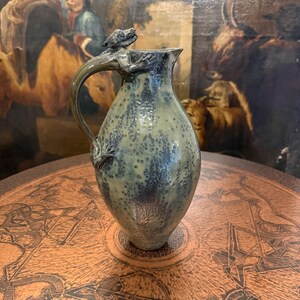 Könnte beinhalten: Ein Keramikkrug mit gesprenkelter grün-dunkelblauer Glasur, der eine skulptierte Eidechsenfigur am Henkel aufweist. Das Gefäß hat einen bauchigen Körper, einen schmalen Hals und einen Ausguss, wobei der Schwanz der Eidechse den Henkel hinunterreicht.