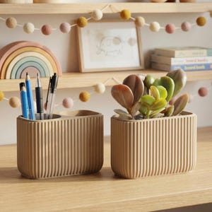 Op de afbeelding: Twee beige, geribbelde bureau-organizers. Eén bevat potloden en pennen, de andere een vetplant. Een houten regenboog en een ingelijste foto staan op een plank erboven. Een slinger van vilten ballen versiert de plank.