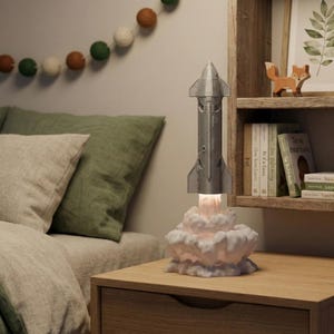 May include: A silver rocket ship lamp with a cloud base on a wooden bedside table. The lamp is beside a bed with green and beige bedding. A decorative garland hangs above the bed. A wooden shelf with books and figurines is in the background.