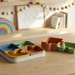May include: A wooden rainbow stacking toy and a multi-colored puzzle tray sit on a wooden surface. The puzzle tray is divided into four sections of blue, yellow, orange, and green. A second puzzle piece is also visible.