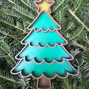 May include: A layered Christmas tree ornament with a green interior and brown outline. A gold star tops the tree. The ornament is set against a backdrop of green pine needles.