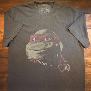 May include: A gray t-shirt featuring a graphic of Raphael from Teenage Mutant Ninja Turtles. The design shows Raphael's face with a red mask. The shirt has a crew neck and short sleeves. The shirt is laid flat on a wooden surface.
