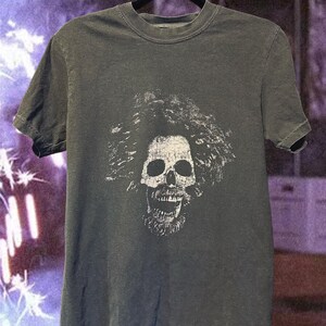 May include: Dark gray t-shirt featuring a white skull graphic with a textured, stylized design. The skull has a detailed beard and hair. The shirt is hanging on a black hanger.