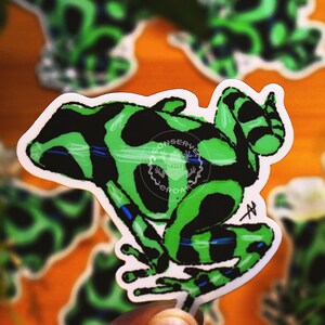 May include: A sticker featuring a green and black poison dart frog design. The frog has a white border and is set against a blurred background of other stickers and foliage. The sticker has the text "CONSERVE" and "WORK" in a circular logo.