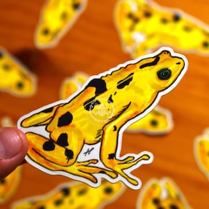 May include: A yellow frog sticker with black spots and green eyes is held in a hand. The sticker has a white border and is set against a blurred background of other frog stickers. The frog is in a side profile pose.