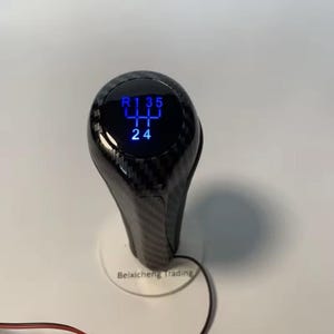 Op de afbeelding: Een koolstofvezel auto versnellingspookknop met een digitale display die de versnellingsposities in blauw weergeeft. De knop heeft een glanzende afwerking en een textuurpatroon. De tekst "R 1 3 5" en "2 4" is zichtbaar.