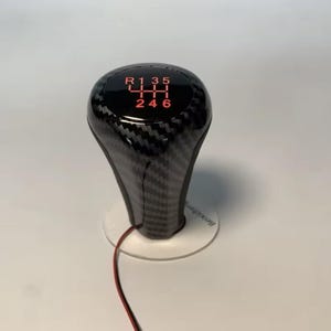Op de afbeelding: Een zwarte en grijze koolstofvezel versnellingspookknop met rood verlichte versnellingsnummers. Het schakelpatroon omvat achteruit, eerste tot en met zesde versnelling. De knop is gemonteerd op een witte basis.