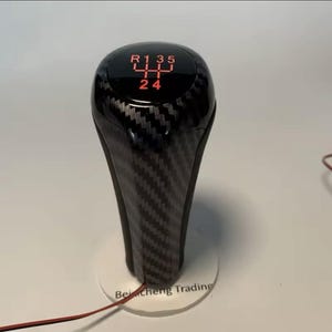 May include: A black and carbon fibre gear shift knob with red illuminated gear numbers. The shift pattern is displayed on the top. The knob is on a white base with the text "BeiCheng Trading".