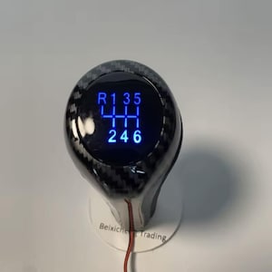 Op de afbeelding: Een zwarte en grijze auto versnellingspookknop met een koolstofvezelpatroon. De knop heeft een blauw verlicht display met de versnellingsposities: R, 1, 3, 5, 4, 2 en 6. De basis van de knop is wit met de tekst "Beixicheng Trading".