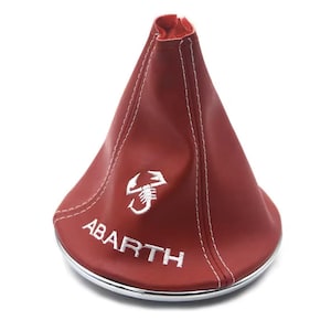 Op de afbeelding: Een rode leren versnellingspookhoes met witte stiksels en een chromen basis. De hoes heeft het wit geborduurde woord "ABARTH" en een schorpioenlogo. Ontworpen voor een auto.