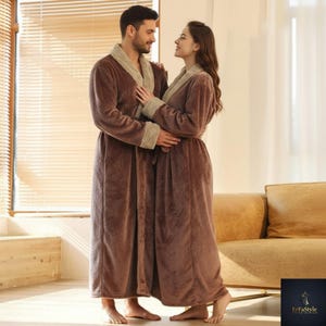 Pode incluir: Duas pessoas vestindo roupões marrons combinando com acabamento marrom claro. Os roupões são longos e têm um cinto. O fundo inclui uma janela com persianas e um sofá bege.