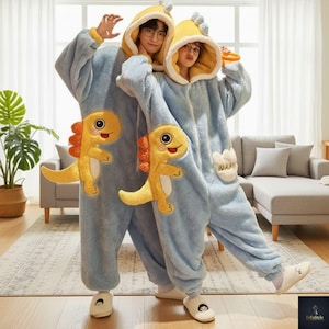 Op de afbeelding: Twee mensen dragen bijpassende lichtblauwe dinosaurus onesie pyjama's met gele capuchons en dinosaurus applicaties. De pyjama's hebben een zachte textuur en zijn aan de voorkant dichtgeritst. Ze staan in een woonkamer.