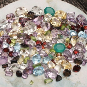 May include: A collection of various gemstones in different shapes and sizes, including amethyst, citrine, aquamarine, and garnet. The gemstones are displayed on a white plate.