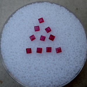 May include: Ten small, square, red gemstones on a white background.