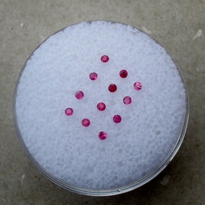 Pode incluir: Uma coleção de doze pequenas pedras preciosas redondas de cor rosa dispostas em forma de diamante sobre um fundo branco.