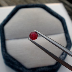 May include: A bright red gemstone held by tweezers. The gemstone is round and appears to be a ruby.
