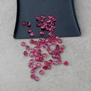 May include: A collection of pink gemstones, likely rubies, scattered on a gray surface. The gemstones are round and have a smooth, polished finish.