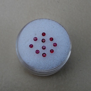 Puede incluir: Un pequeño recipiente blanco con tapa, lleno de 12 pequeñas piedras preciosas redondas de color rojo.