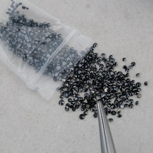 May include: A plastic bag filled with loose black diamonds, some of which have spilled out onto a gray surface. A pair of tweezers is visible in the lower right corner of the image.