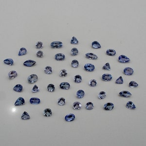 May include: An assortment of blue gemstones in various shapes, including oval, pear, and round cuts. The gemstones are scattered across a white surface, showcasing their facets and color variations. The gems are likely for jewelry making.