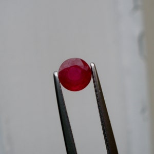 May include: A vibrant red gemstone held by tweezers. The gemstone is oval-shaped and appears to be a ruby.
