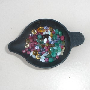 May include: A black metal dish filled with a variety of colorful gemstones, including green, red, blue, yellow, and white.