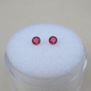 Puede incluir: Un par de pequeñas piedras preciosas rojas, redondas y facetadas, con talla brillante. Las dos piedras circulares están colocadas una al lado de la otra, mostrando un tono carmesí profundo y superficies reflectantes sobre una base de espuma.