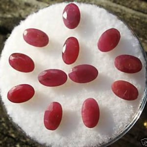 May include: Ten oval-shaped, faceted ruby gemstones in various shades of red. The gemstones are arranged in a circular pattern on a white background.