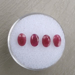 May include: Four oval-shaped ruby gemstones, each with a deep red color, are displayed in a row on a white background.