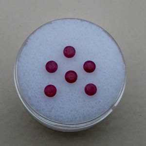 May include: Six round, red gemstones in a clear plastic container with a white background.