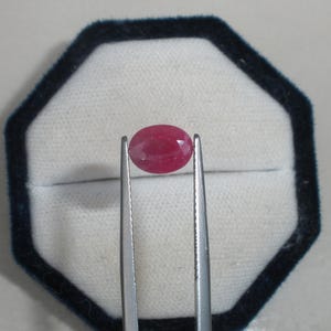 May include: A vibrant, oval-cut ruby gemstone with a deep pinkish-red hue and faceted surface, held securely by a pair of silver-toned metal tweezers. The gemstone displays a translucent quality and is positioned centrally against a white background.