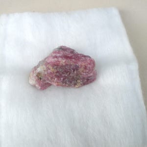 Peut inclure: Une pierre précieuse naturelle brute avec un mélange de teintes roses et blanches. La pierre a une surface texturée et est posée sur un fond blanc et duveteux. Il s'agit d'un spécimen minéral.