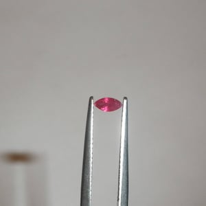 May include: A close-up of a bright pink gemstone held by tweezers against a white background.