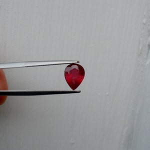 Pode incluir: Uma gema de cor vermelha profunda em forma de pêra, segurada por pinças contra um fundo branco.