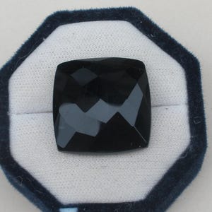 May include: A square-shaped black gemstone with a faceted surface, featuring a cushion cut. The stone is displayed centred within an octagonal jewellery box that has a dark blue velvet border and a white interior lining for contrast.