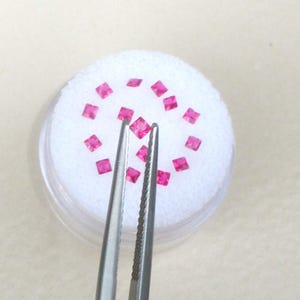 Op de afbeelding: Een verzameling kleine, vierkante, felroze edelstenen, tentoongesteld op een witte ondergrond in een transparante container. Een zilveren pincet houdt een van de edelstenen vast. De edelstenen zijn waarschijnlijk voor sieraden.