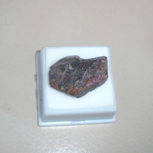 May include: A dark red and brown gemstone with a rough, irregular shape, displayed in a white square box.