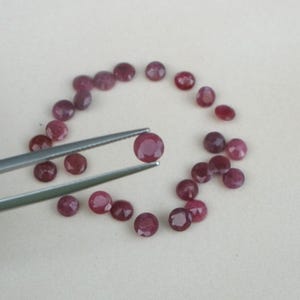 May include: A collection of small, round, faceted rubies in varying shades of red and purple. One ruby is held by silver tweezers, with the others arranged in a loose circle. The gemstones are displayed on a light-colored surface.